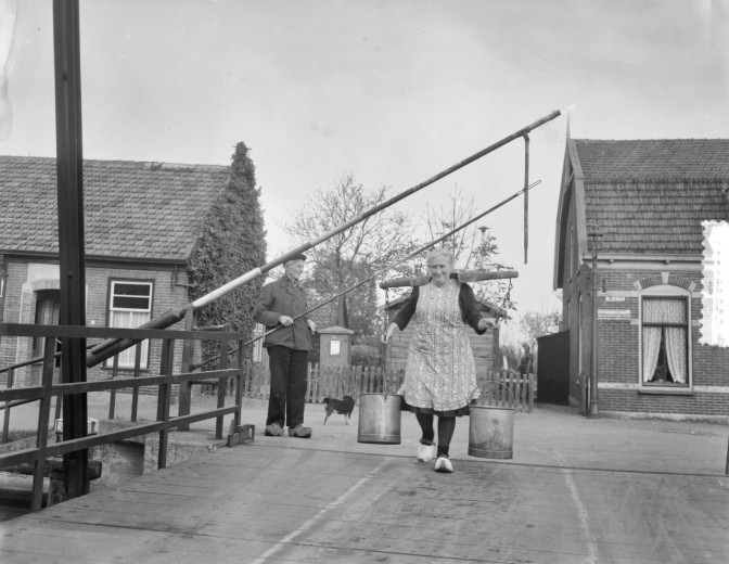 woman carrying milk on a yoke over her shoulders across a toll bridge